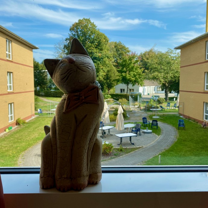 Eine Katze aus Keramik gefertigt, steht auf eine Fensterbank. Der Blick fällt in den dahinterliegende Garten mit Sonnenschirmen und Gartenmöbeln (vergrößerte Bildansicht wird geöffnet)