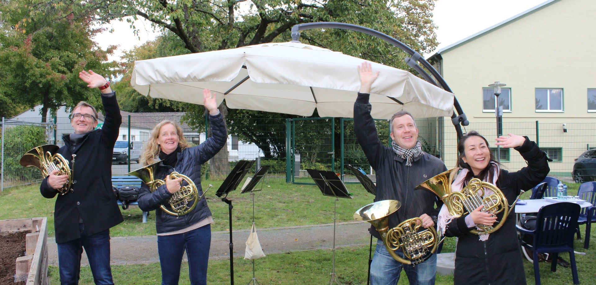 Drei Männer und ein Frau halten Horninstrumente in ihren Händen und winken