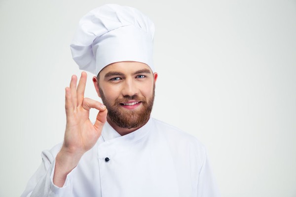 Ein bärtiger Mann mit Kochmütze und weißer Kochjacke formt Daumen und Zeigefinger zu einem O