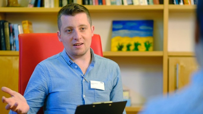 Ein junger Mann mit LWL-Namensschilkd sitz vor einem Bücherregal