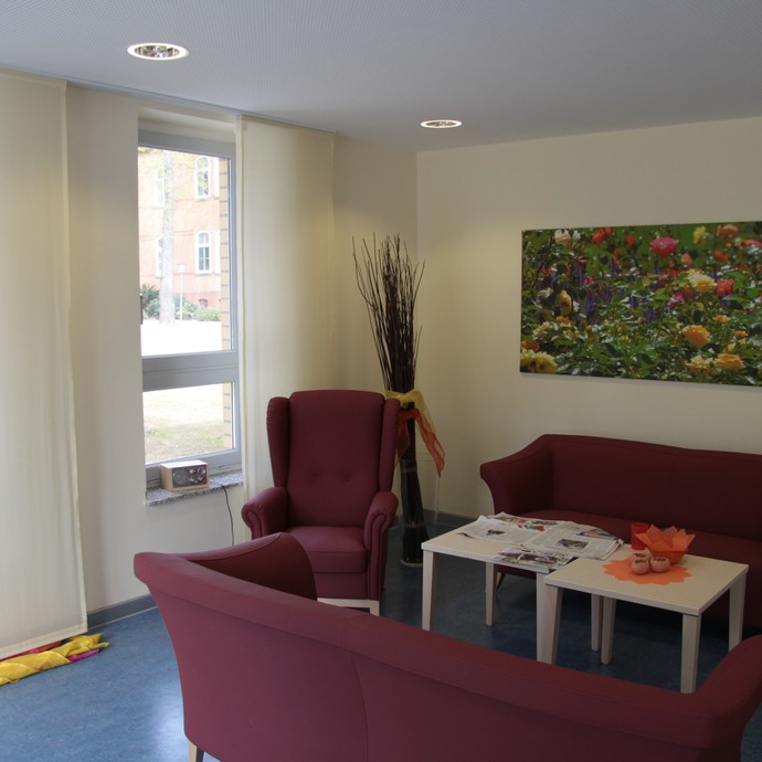 Blick in ein Wohnzimmer mit weinroter Couch und einem Ohrensessel. Dahinter ein farbenfrohes Bild einer Blumenwiese (öffnet vergrößerte Bildansicht)